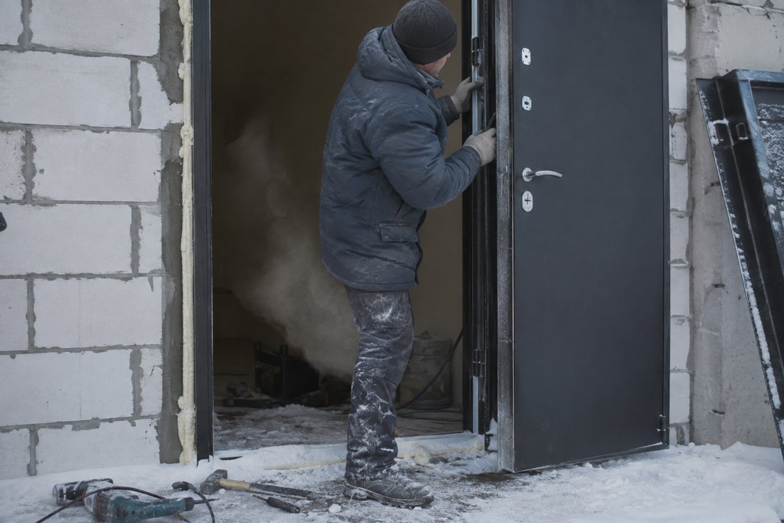 Мастер устанавливает металлическую входную дверь зимой, на пороге снег, из открытого проёма идёт пар